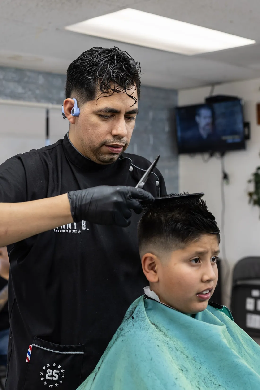 A barber combs and trims the top of a young boy's haircut while the boy sits in a teal cape