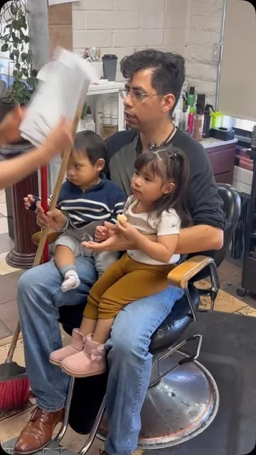 A father sits in a barber chair holding his two toddlers on his lap at Ema's Family Barbershop