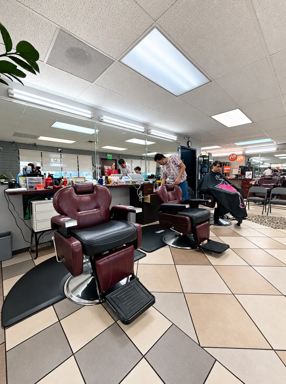 Bright interior of Ema's Family Barbershop with burgundy barber chairs on a checkered tile floor, mirrored stations and ceiling lights