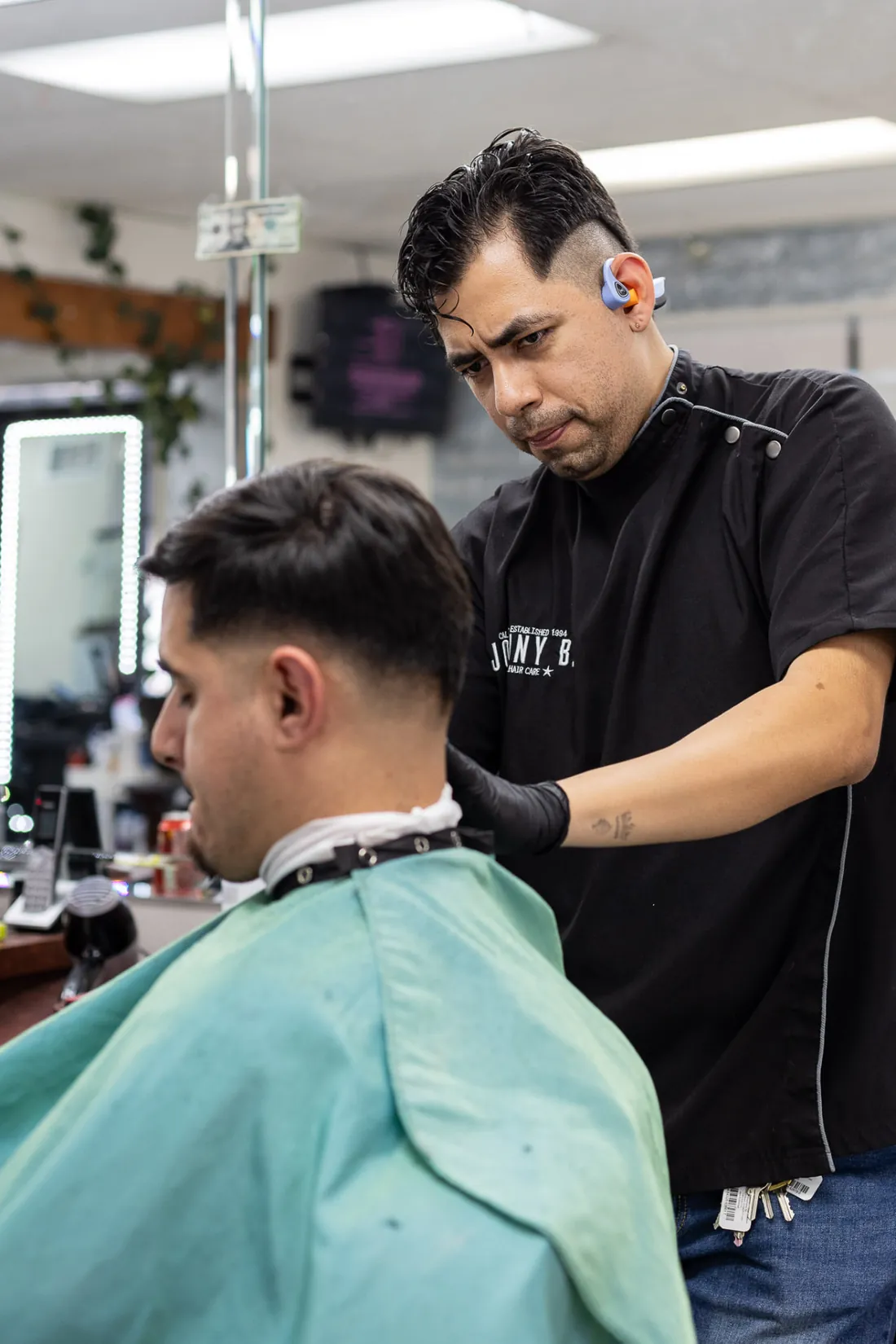 A barber in a black shirt and gloves shapes a client's fade at a station inside the shop